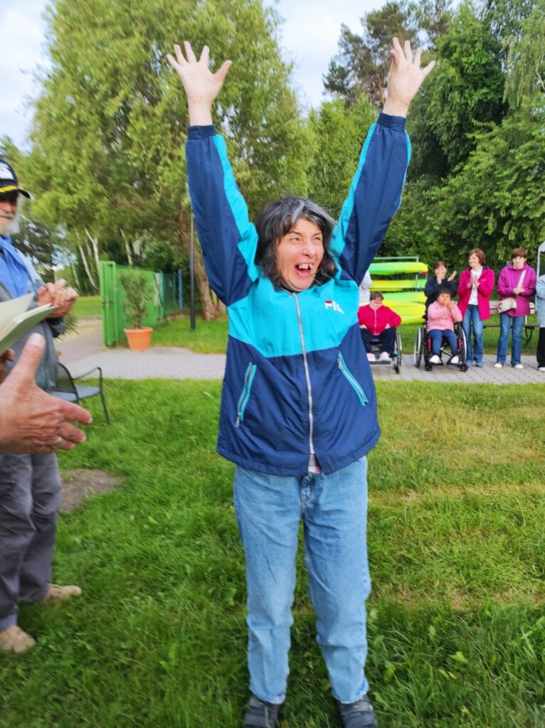 Kobieta z podniesionymi rękami stojąca na trawniku a w tle siedzące osoby na  krzesłach
