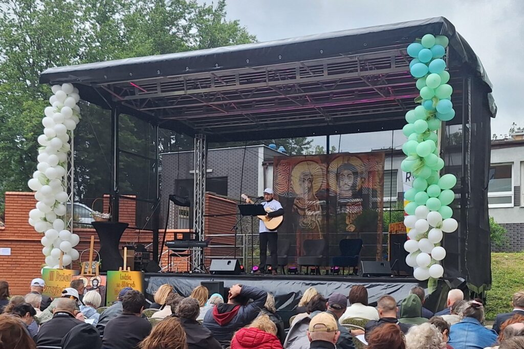 Mężczyzna z gitarą stojący na plenerowej scenie ozdobionej białymi i zielonymi balonami