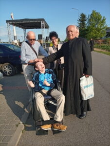 Na asfaltowej drodze trzy dorosłe osoby stojące przy osobie siedzącej na wózku inwalidzkim