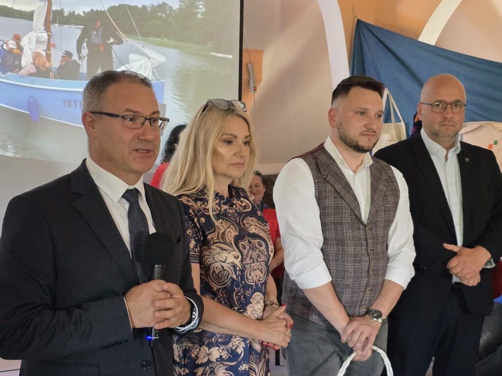 Trzech mężczyzn i kobieta stojących na tle wyświetlanego zdjęcia z płynącą żaglówką