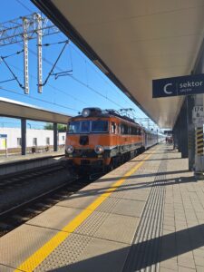 Wjeżdżający na peron pociąg pasażerski z pomarańczową lokomotywą i szarymi wagonami