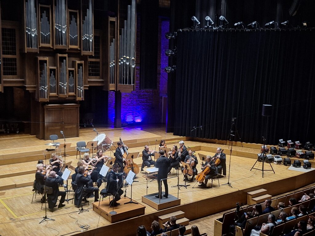 Dyrygent oraz kilkunastoosobowa grupa osób z instrumentami smyczkowymi grająca i siedząca na krzesłach ustawionych na scenie filharmonii