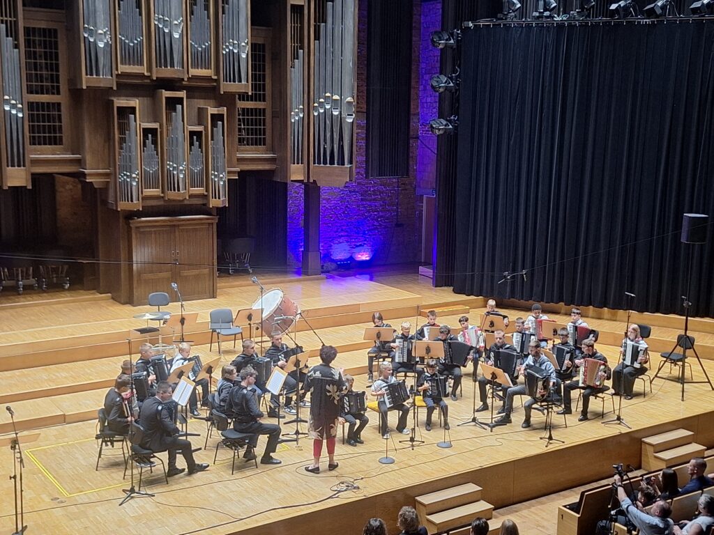 Kilkunastoosobowa grupa młodzieży z akordeonami siedząca na krzesłach ustawionych na scenie filharmonii