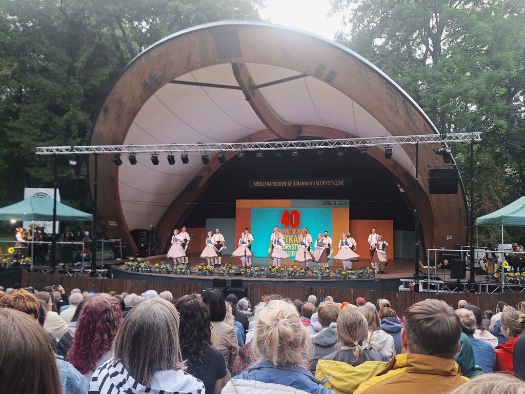 Muszla koncertowa w parku ze sceną na której grupa osób w strojach ludowych tańczy w parach