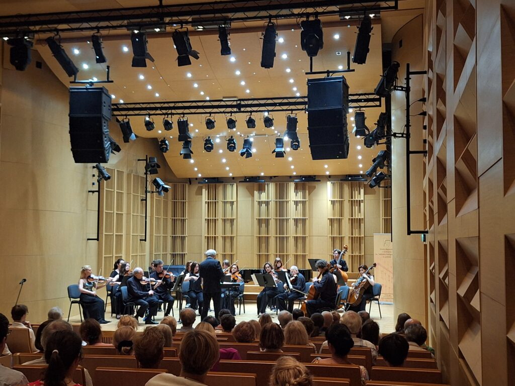 Wnętrze sali koncertowej z siedzącą publicznością a w tle dyrygent oraz grupa muzyków grających na instrumentach smyczkowych