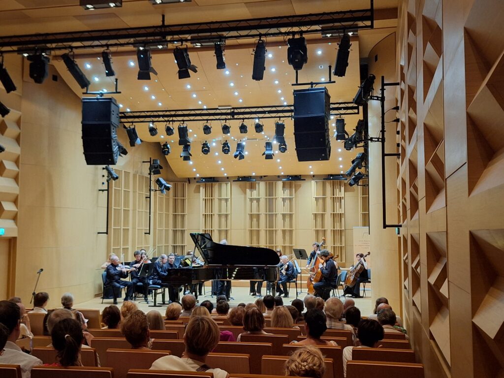 Wnętrze sali koncertowej z siedzącą publicznością a w tle na scenie pianista przy fortepianie oraz grupa muzyków grających na instrumentach smyczkowych