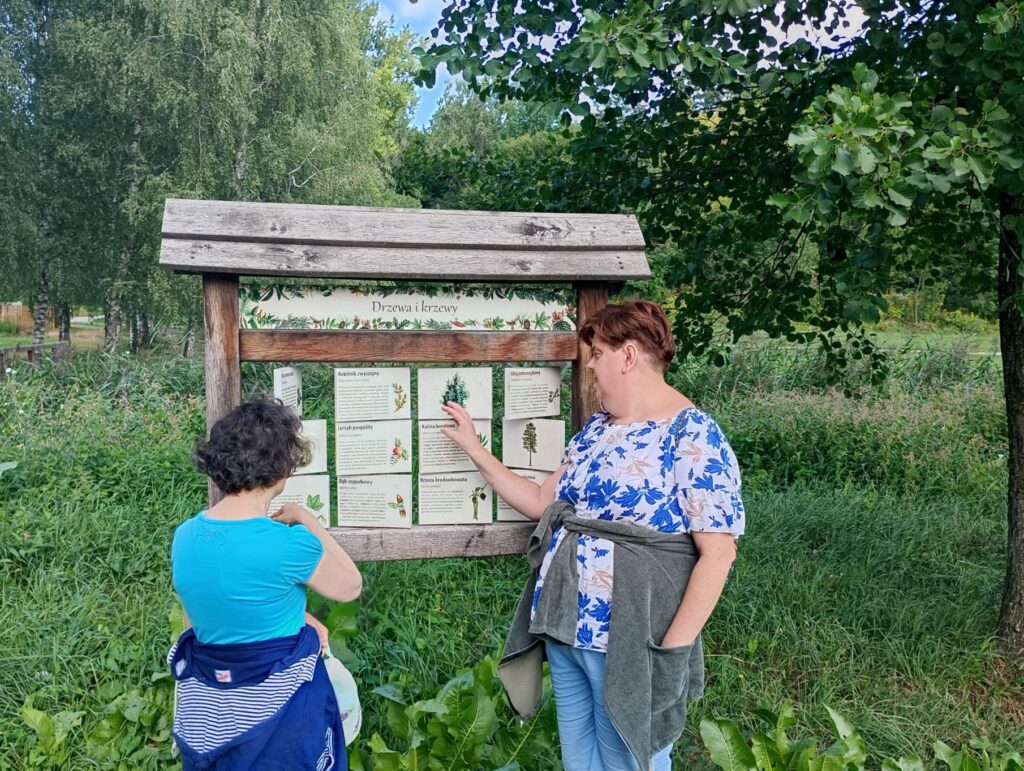 Dwie kobiety stojące przy tablicach edukacyjnych z wizerunkami i nazwami drzew oraz krzewów