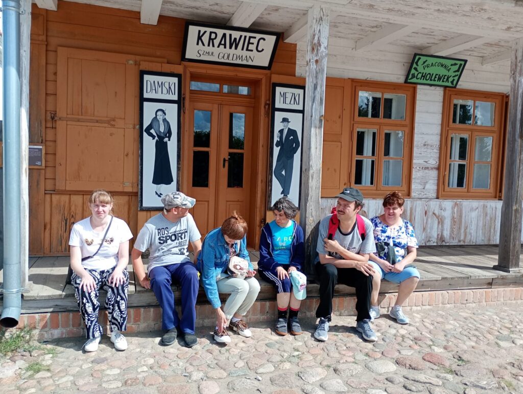 Sześć dorosłych osób siedzących przed wejściem do zakładu krawieckiego drewnianej zabudowy rynkowej w skansenie