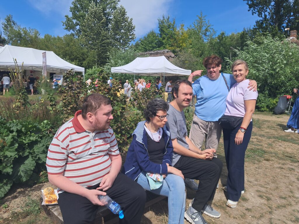 Dwie kobiety i obok trzy siedzące osoby na drewnianym balu w otoczeniu zieleni z widocznymi białymi namiotami w tle
