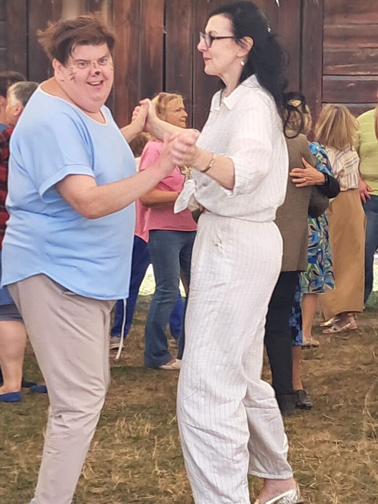 Dwie kobiety tańczące w parze i inne pary widoczne po bokach na tle ściany z ciemnobrązowych desek