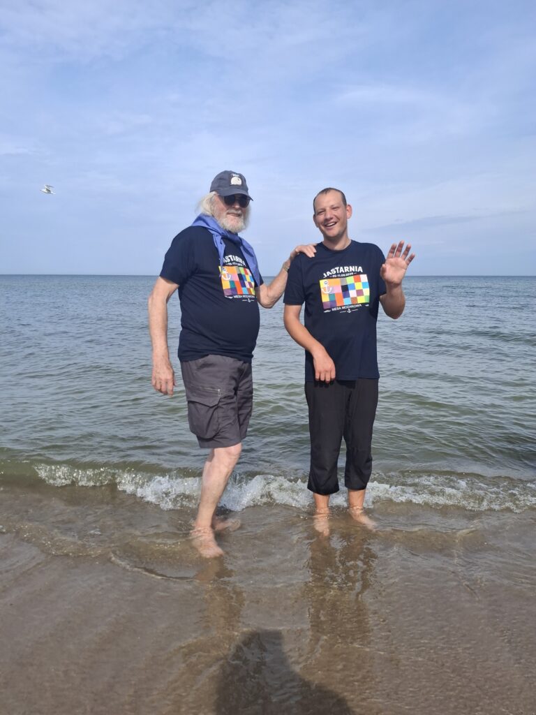 Dwóch mężczyzn w krótkich spodenkach stojących bosymi stopami w morzu i patrzących w stronę widza