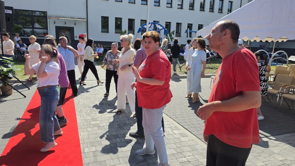 Grupa dorosłych osób stojąca w rzędach na chodniku przed białym budynkiem