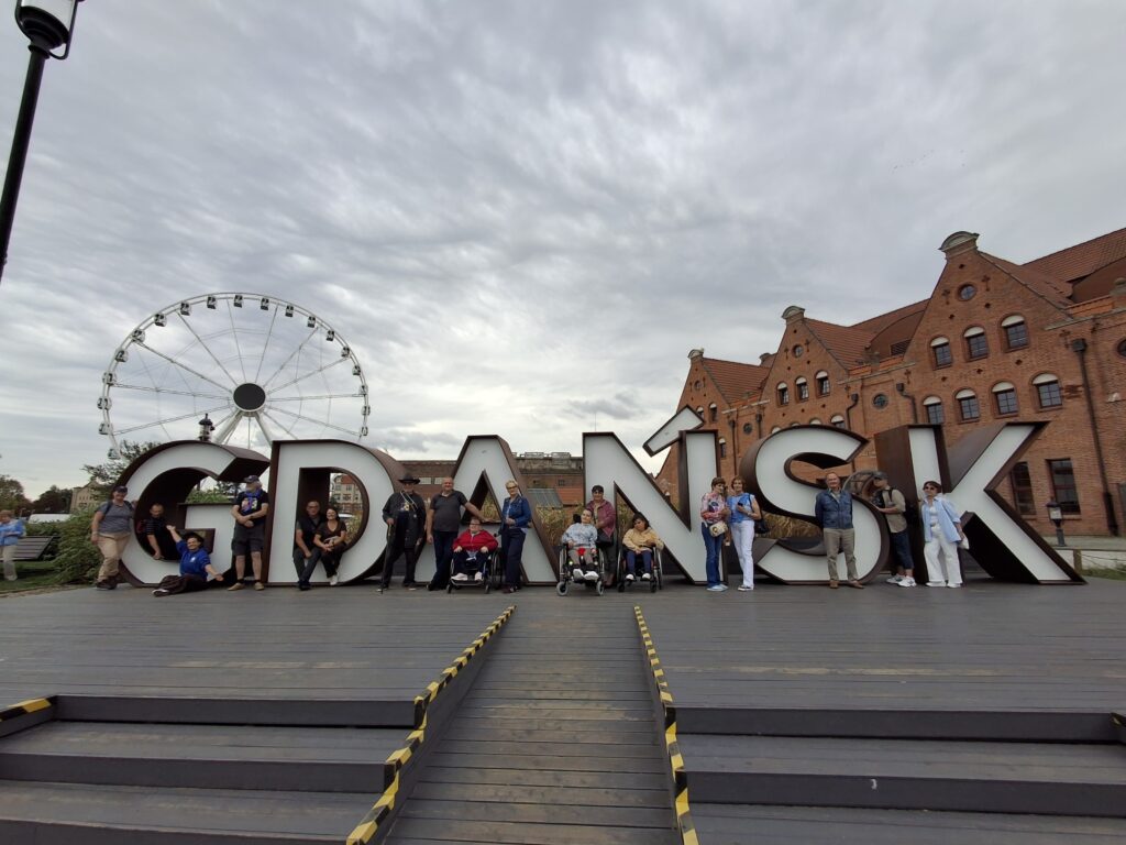 Grupa dorosłych osób stojących pomiędzy białymi literami GDAŃSK na tle kamienic z czerwonej cegły oraz koła widokowego