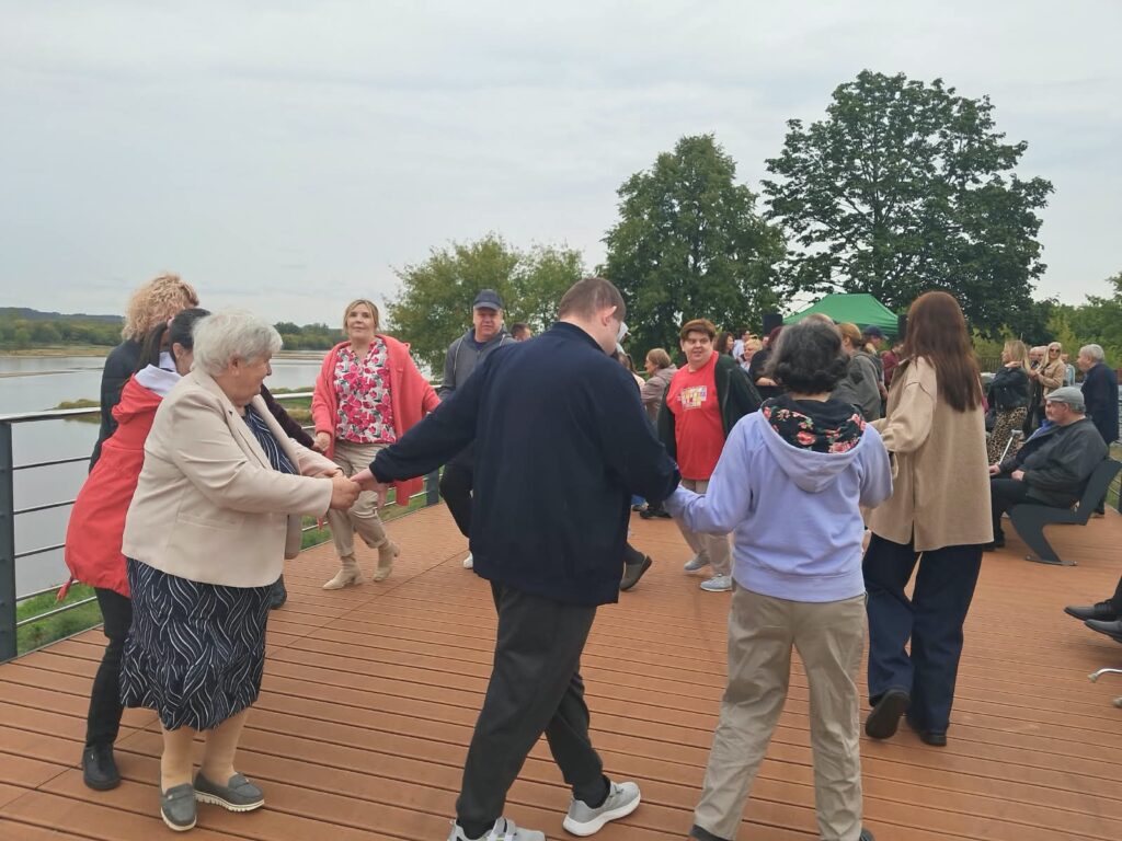 Grupa dorosłych osób tańczących w kręgu na pomoście przy rzece płynącej po lewej stronie