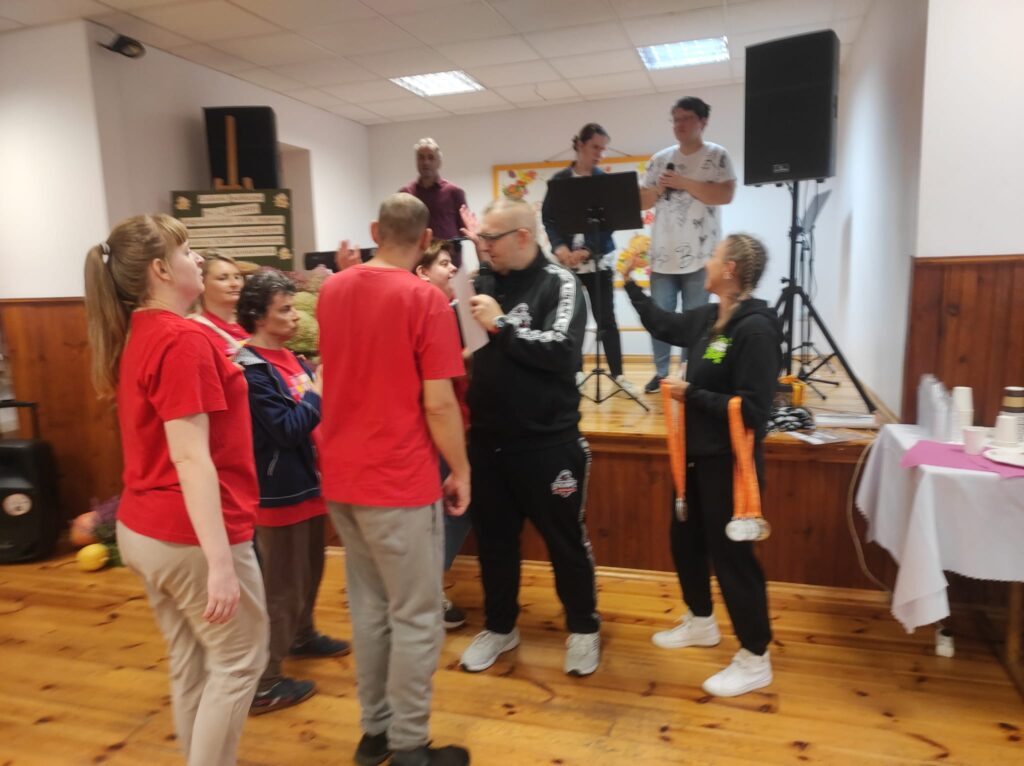 Grupa dorosłych osób we wnętrzu przed sceną oraz na scenie podczas zabawy tanecznej