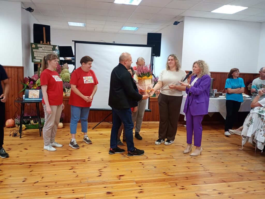 Grupa osób stojących w sali na drewnianej podłodze na tle białego ekranu projekcyjnego