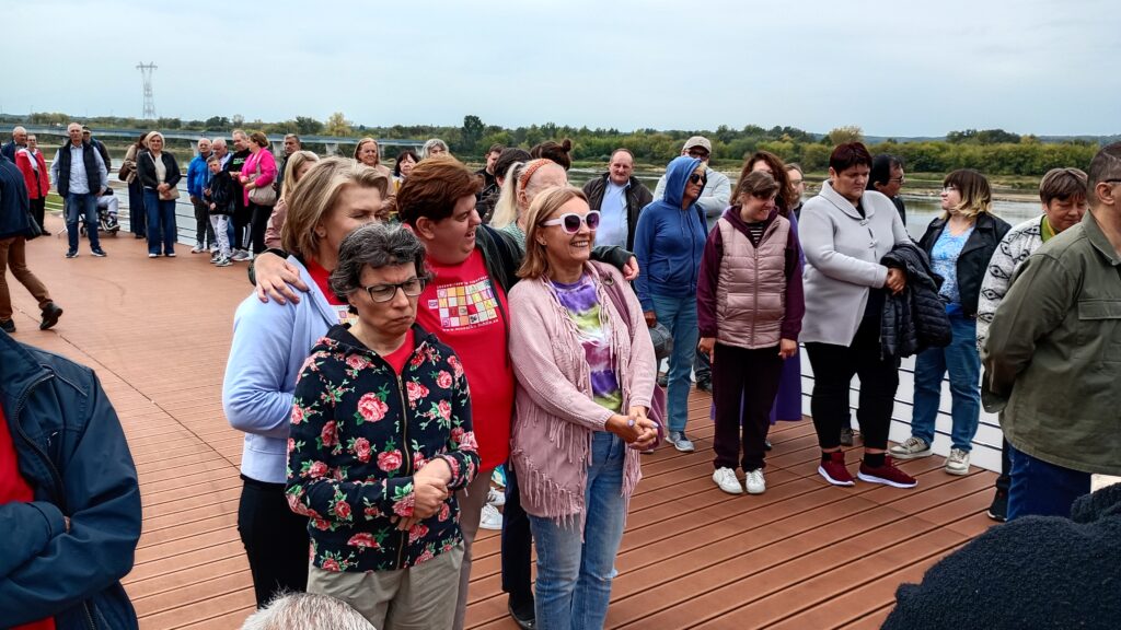 Kilka grup dorosłych osób stojących na pomoście przy rzece płynącej po prawej stronie