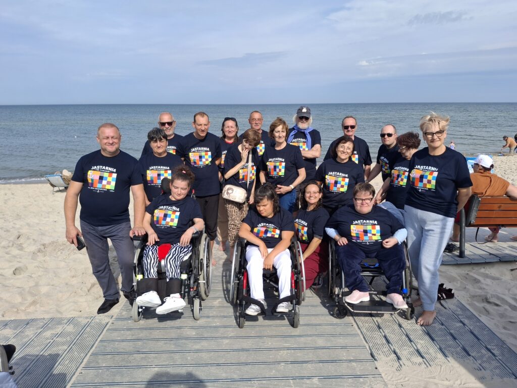 Kilkunastoosobowa grupa dorosłych osób w czarnych podkoszulkach z logo stojąca na plaży na tle morza