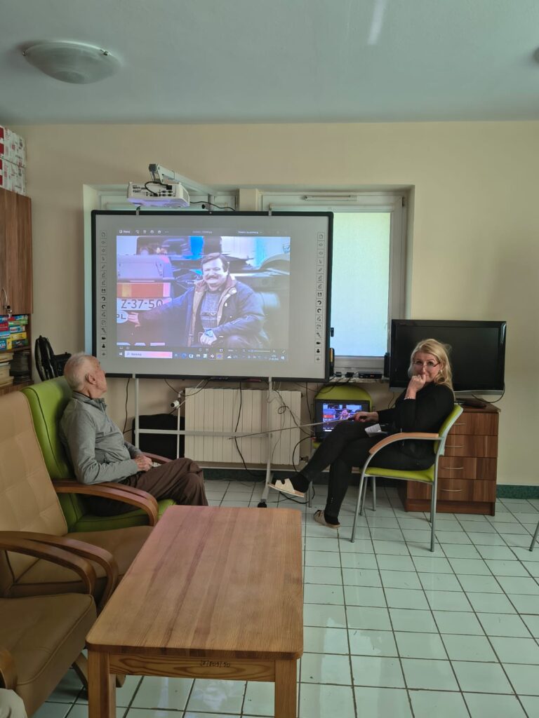 Mężczyzna i kobieta siedzący na zielonych krzesłach ustawionych przed ekranem projekcyjnym na którym widać fotografię mężczyzny przy ciężarówce