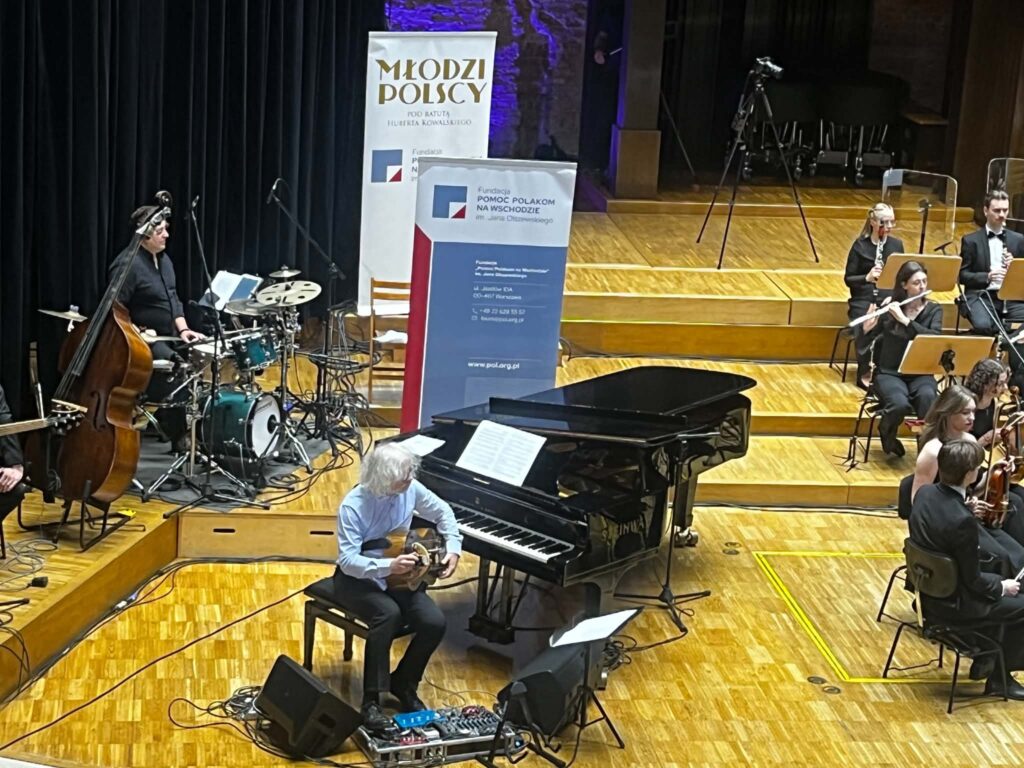 Mężczyzna siedzący z lirą korbową w sąsiedztwie fortepianu  i w otoczeniu członków orkiestry z instrumentami we wnętrzu filharmonii
