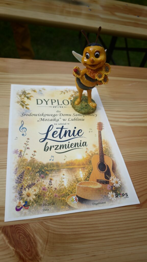Okolicznościowy dyplom leżący na drewnianym blacie oraz kolorowa figurka pszczoły stojąca na nim