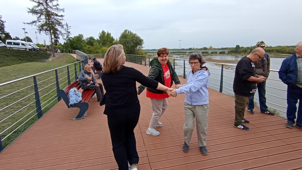Trzy kobiety tańczące w kręgu na pomoście przy rzece płynącej po prawej stronie