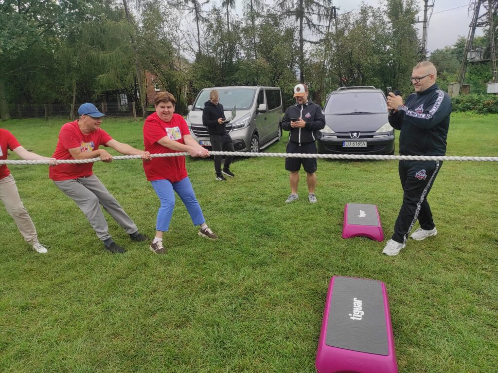 Trzy osoby w czerwonych podkoszulkach stojące na trawie i przechylone na lewą stronę oraz trzymające linę w dłoniach