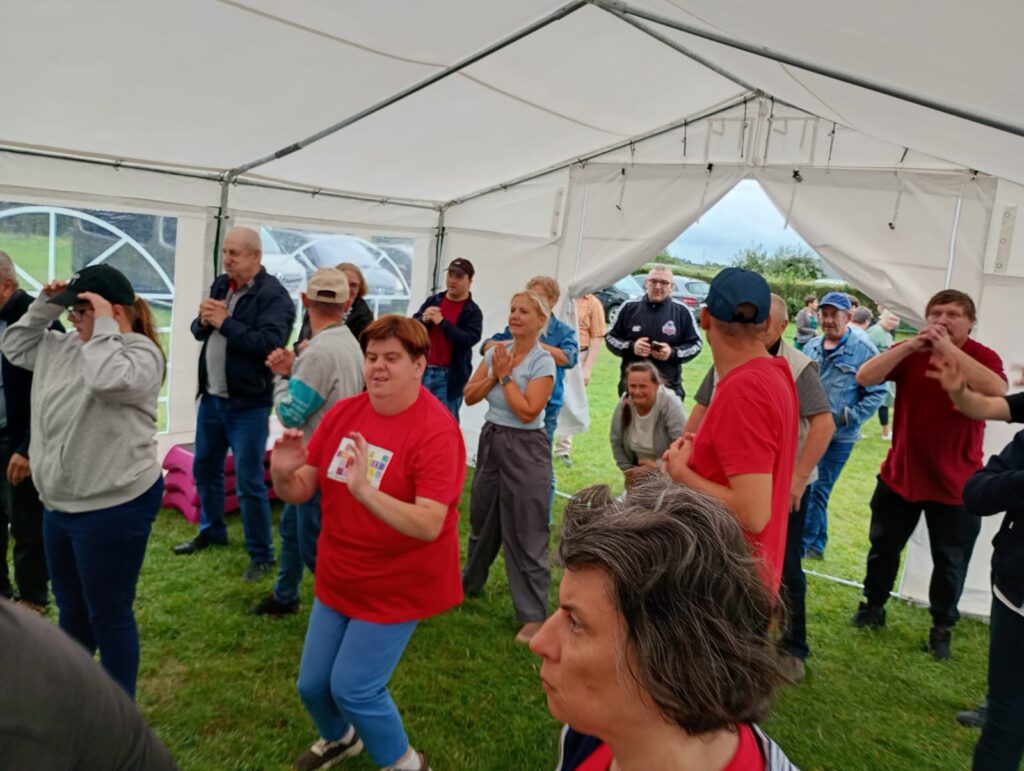 W białym namiocie plenerowym grupa dorosłych osób stojących na trawie i wykonujących ćwiczenia rozgrzewające