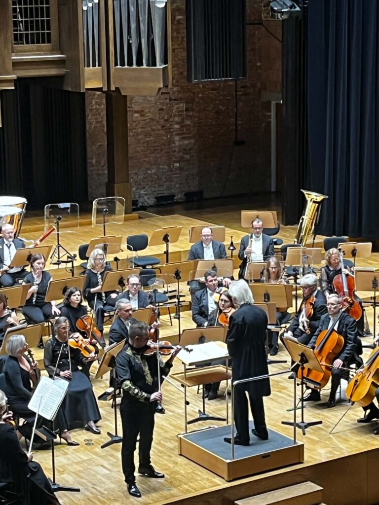 We wnętrzu grupa siedzących dorosłych osób z instrumentami oraz stojący przed nimi mężczyzna z batutą i mężczyzna ze skrzypcami