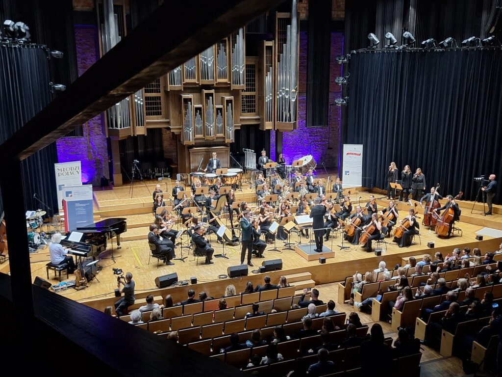 Wnętrze sali w filharmonii z członkami orkiestry z instrumentami oraz widownią wypełnioną publicznością