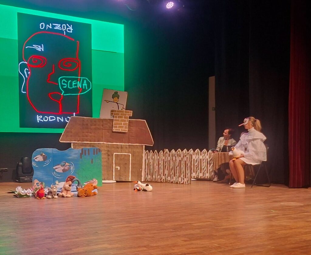 Dwie dorosłe osoby siedzące na scenie obok scenografii imitującej wiejską chałupę z płotem oraz staw z kaczkami