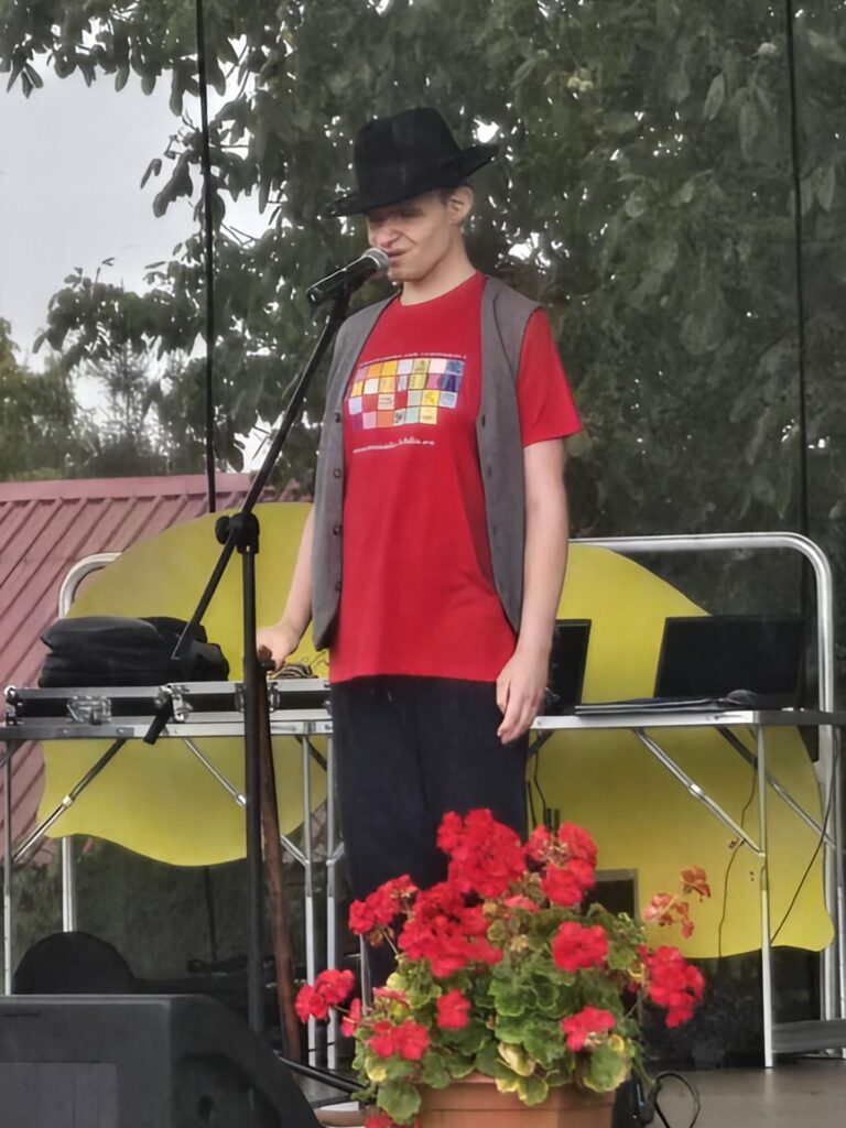 Mężczyzna w czerwonym podkoszulku i czarnym kapeluszu stojący na scenie plenerowej a przed nim czerwone kwiaty w dużej doniczce