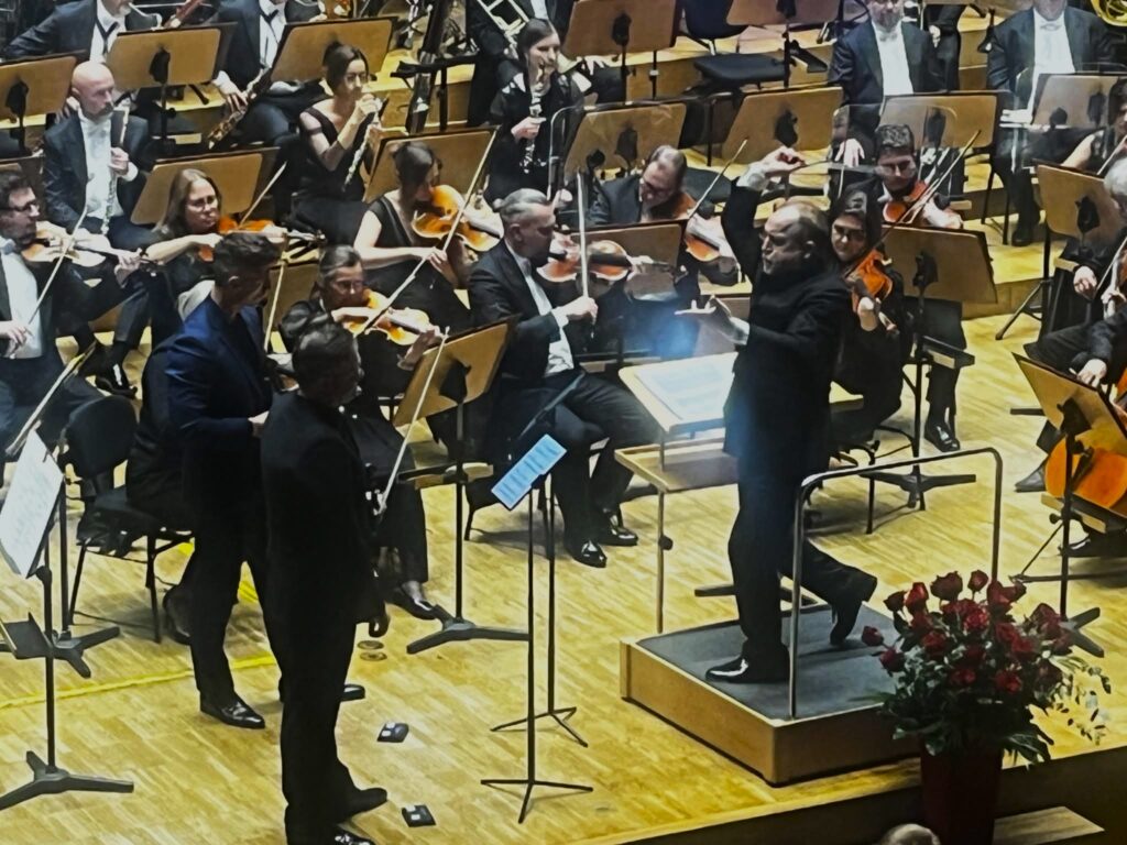 Postać mężczyzny ubranego w garnitur i trzymającego batutę w prawej dłoni stojącego na tle muzyków z instrumentami będących częścią orkiestry