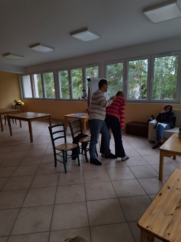 Stojący mężczyzna opierający się o plecy nachylonej kobiety w sali z wieloma oknami